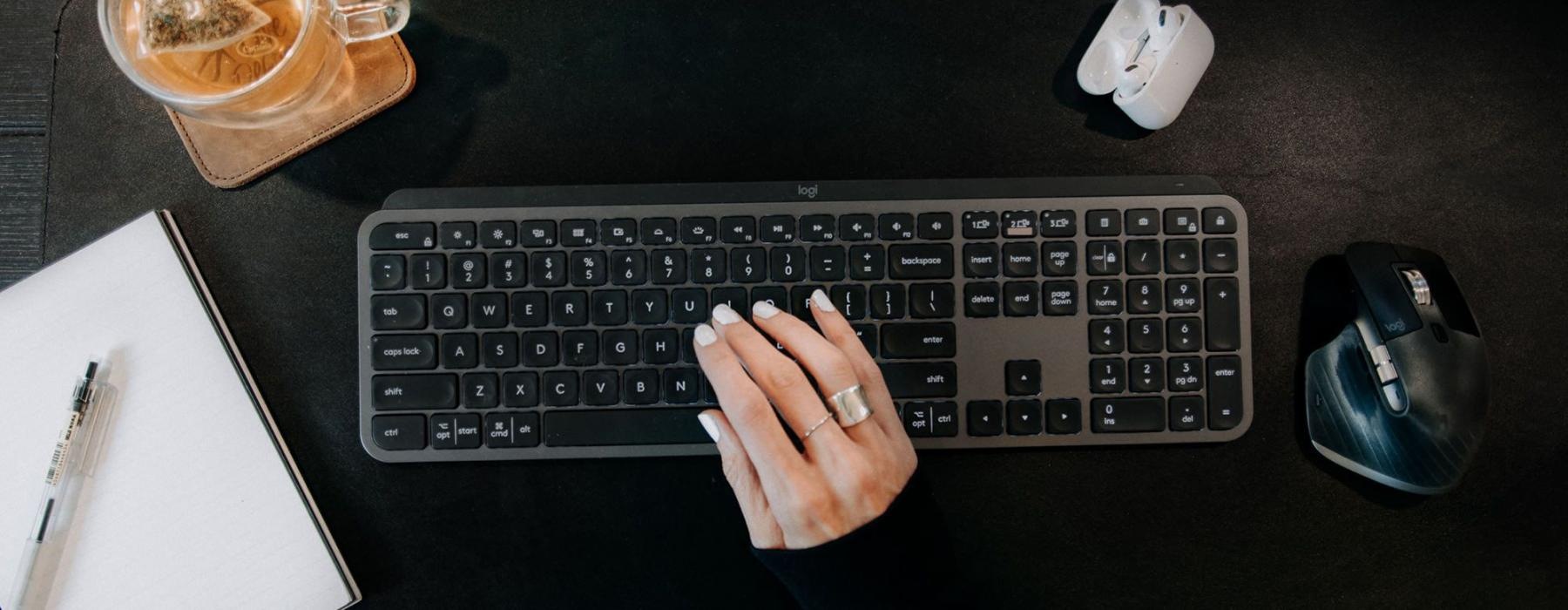 woman's hand on a keyboard surrounded by office items and a cup of tea on a coaster